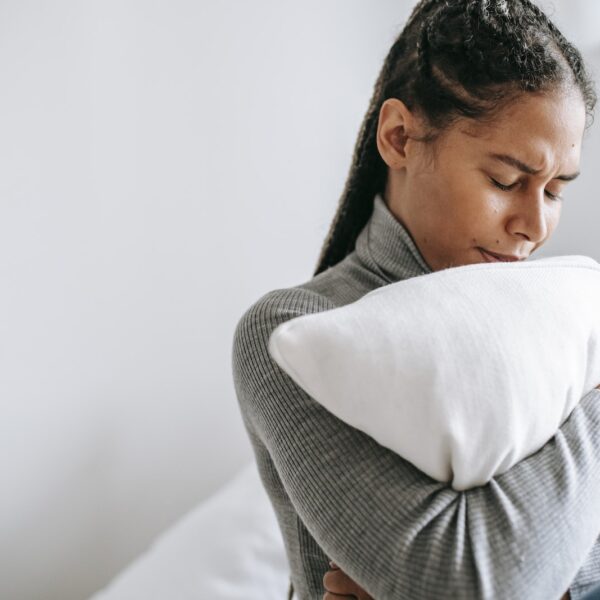 Young ethnic female with closed eyes and sad face expression sitting on bed and embracing pillow